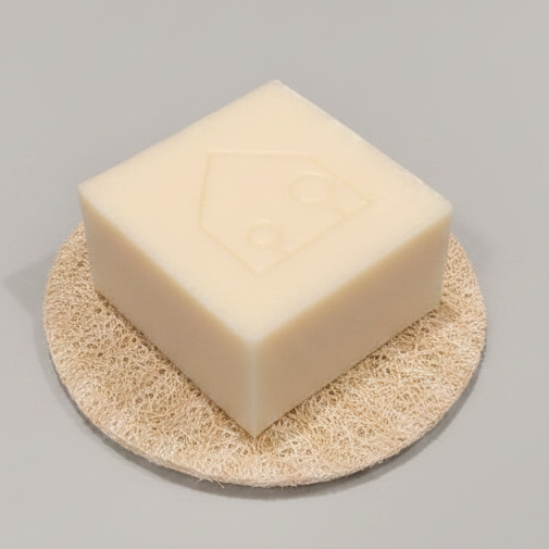A square cream-colored soap bar with an embossed house logo, resting on a round natural loofah sponge against a neutral gray background.