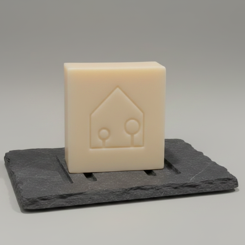 A square cream-colored soap bar with an embossed house and tree logo, displayed on a dark gray slate soap dish against a neutral gray background.