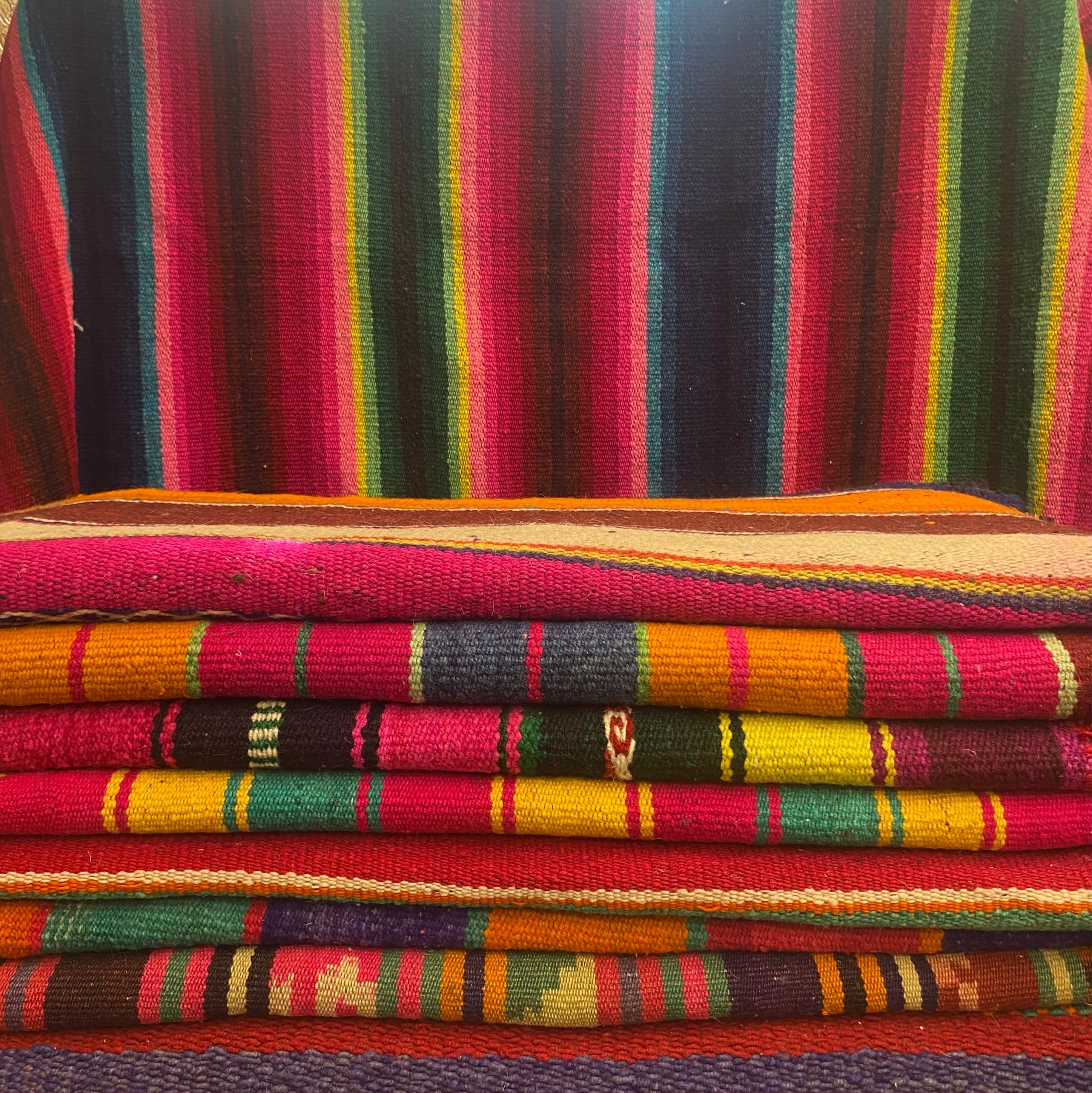 A stack of colorful woven rugs, folded serape-style rugs, in vivid red, pink, green, yellow, orange, and blue tones.