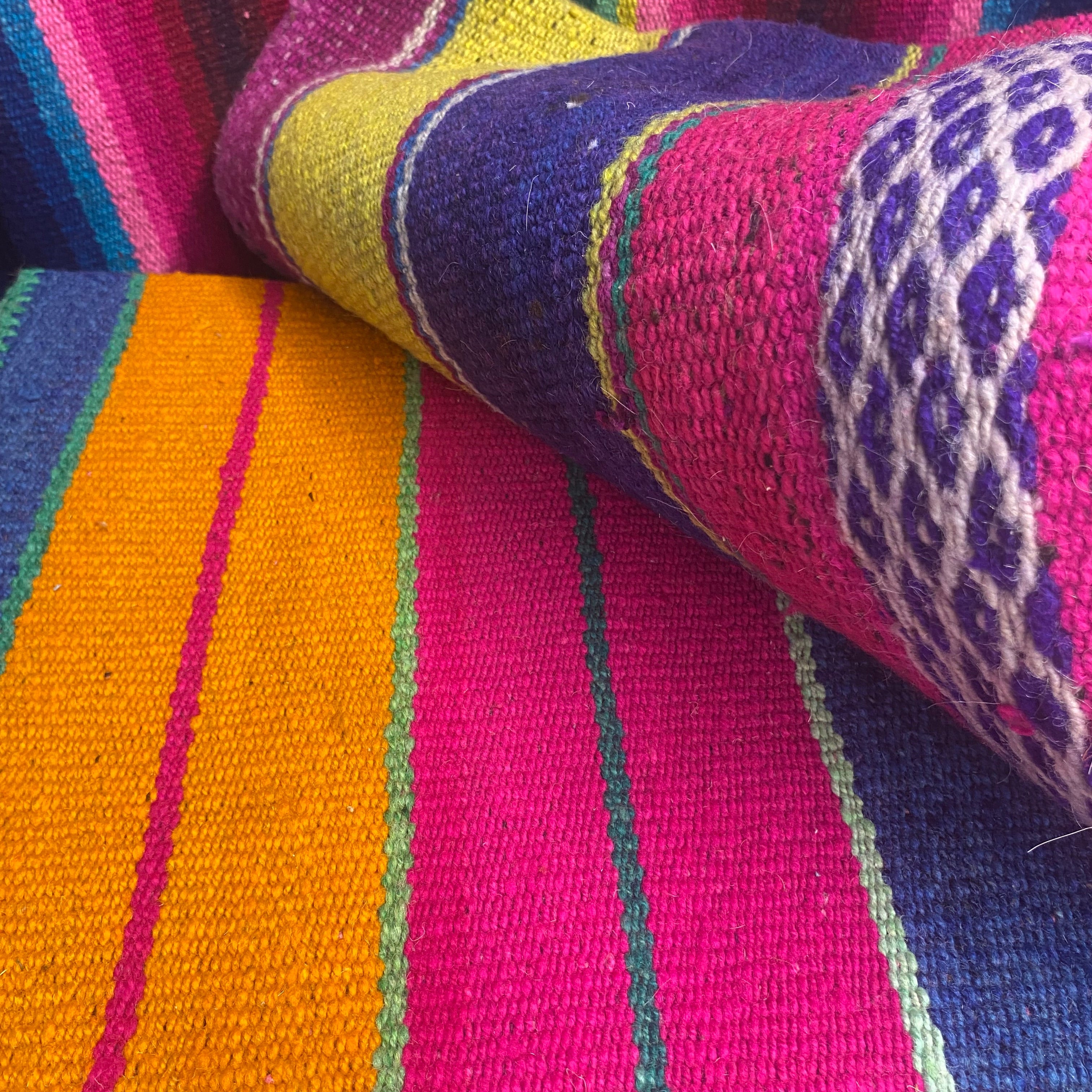 A close-up of a colorful woven rug with bold vertical stripes in pink, orange, purple, blue, green, and yellow, with one corner folded over to show the woven texture and patterned section.