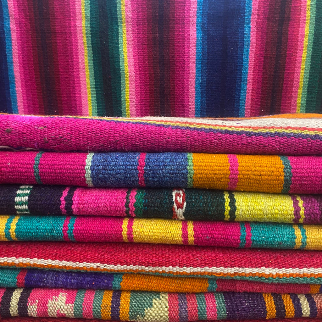 A stack of folded colorful woven rugs with vertical stripes in pink, blue, green, yellow, orange, and purple, with a larger matching striped fabric hanging behind them.