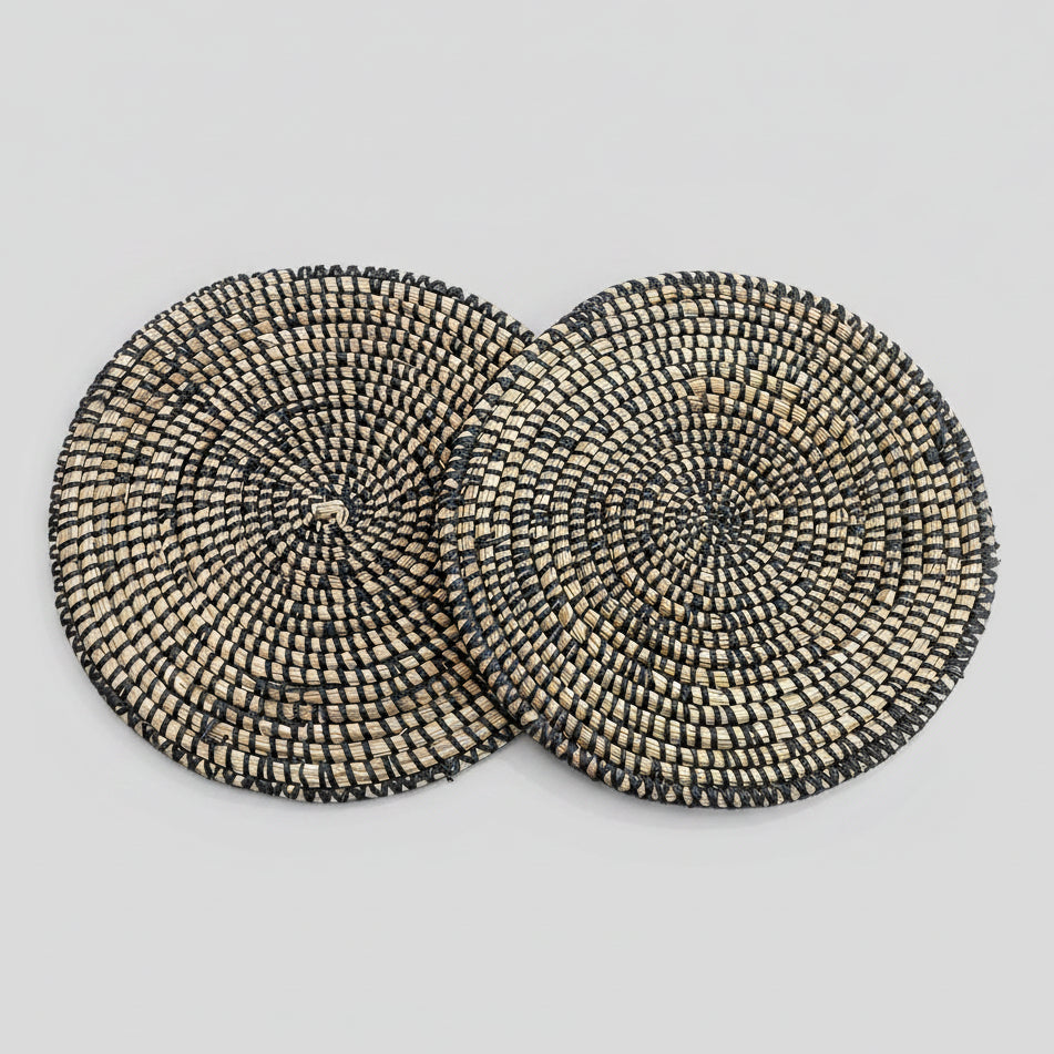 A set of two round placemats with a geometric pattern, featuring a mix of black and beige river grass with white binding.