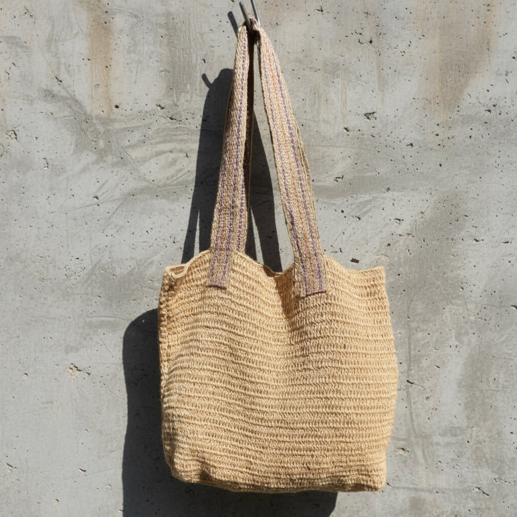 A natural beige woven tote bag with dual shoulder straps, hanging against a textured concrete wall in outdoor sunlight.