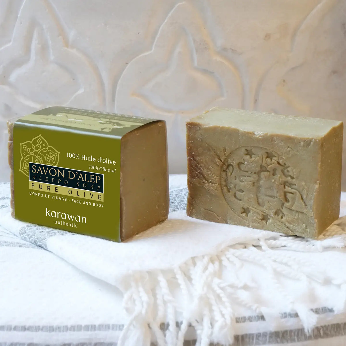 A bar of Karawan Moroccan olive oil soap shown next to its green and gold packaging box, resting on a white fringed textile against a white patterned background.