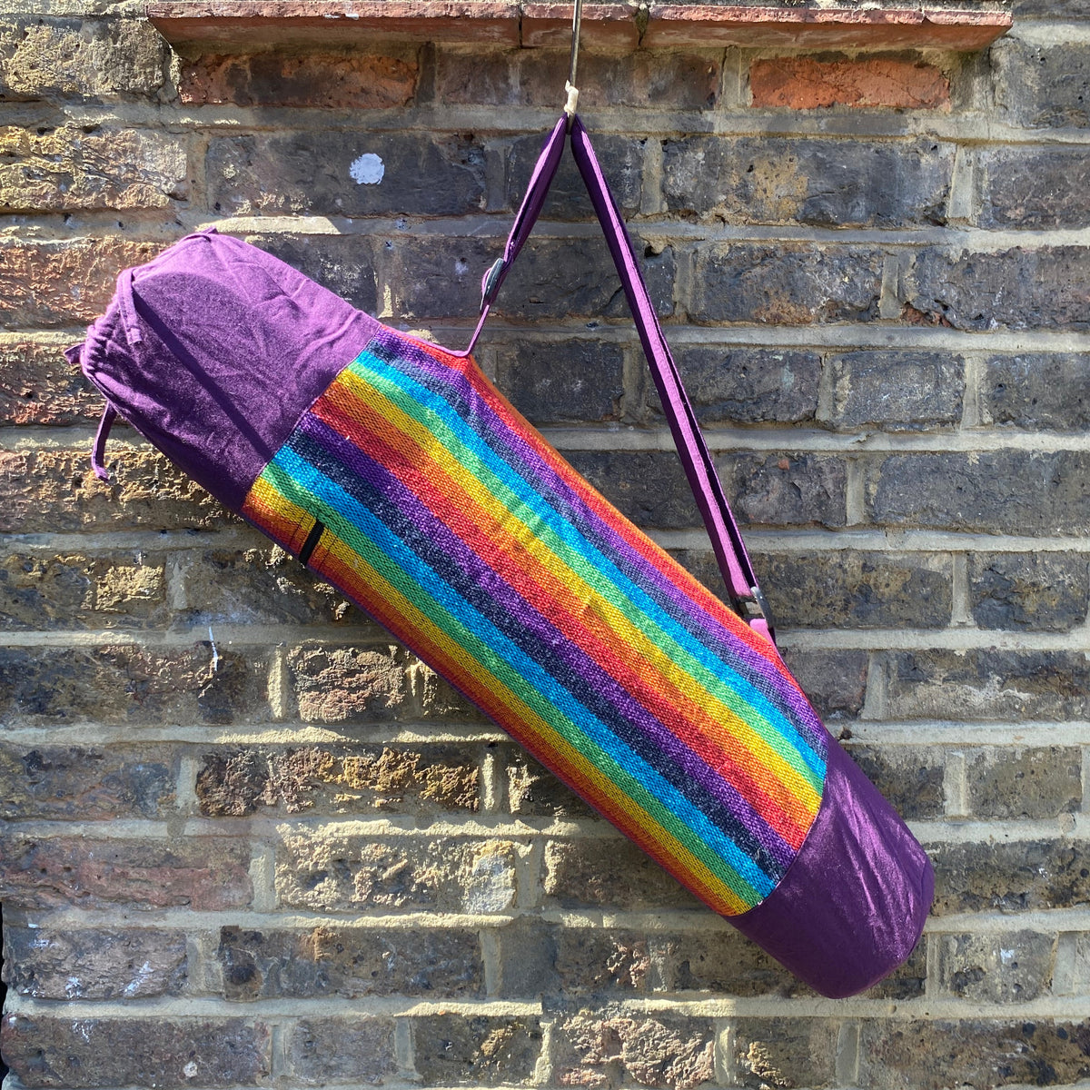 A purple yoga mat roll bag with a vibrant rainbow striped fabric, hanging from a metal hook on a weathered brick wall.