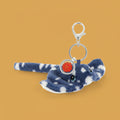 A plush keychain , a blue and white stingray, featuring a silver metal clasp, key ring, and a round red charm with a gold emblem.
