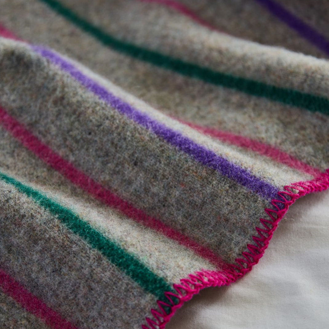 A close-up of a textured wool blanket with a heather gray base and diagonal stripes in purple, green, and pink, finished with a bright pink braided edge.