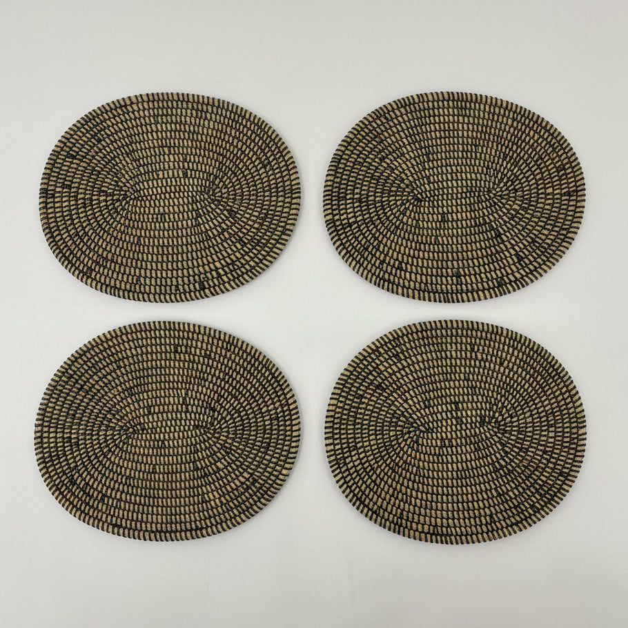 A oval woven placemat made of natural tan and black raffia or seagrass, photographed on a plain white fabric background.