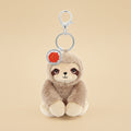 A plush sloth keychain charm with a silver metal clasp and key ring, featuring a light brown and cream sloth face with a small red circular tag.