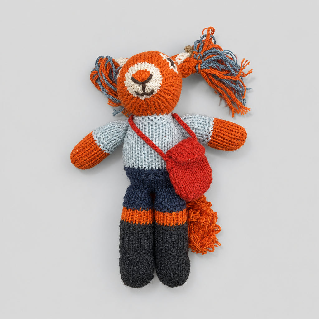 Hand-knitted red squirrel soft toy wearing a grey top, dark shorts and a red shoulder bag.”