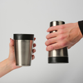 Two hands holding stainless steel travel tumblers with black lids against a light gray background.