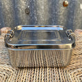 A rectangular stainless steel lunch box with a fitted lid and loop handles, placed on a woven wicker surface against a corrugated metal wall.