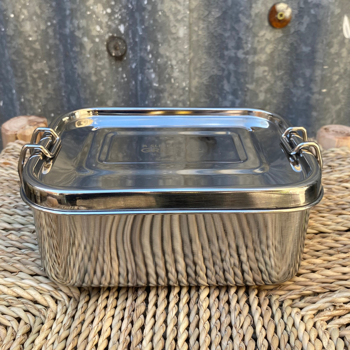 A rectangular stainless steel lunch box with a fitted lid and loop handles, placed on a woven wicker surface against a corrugated metal wall.