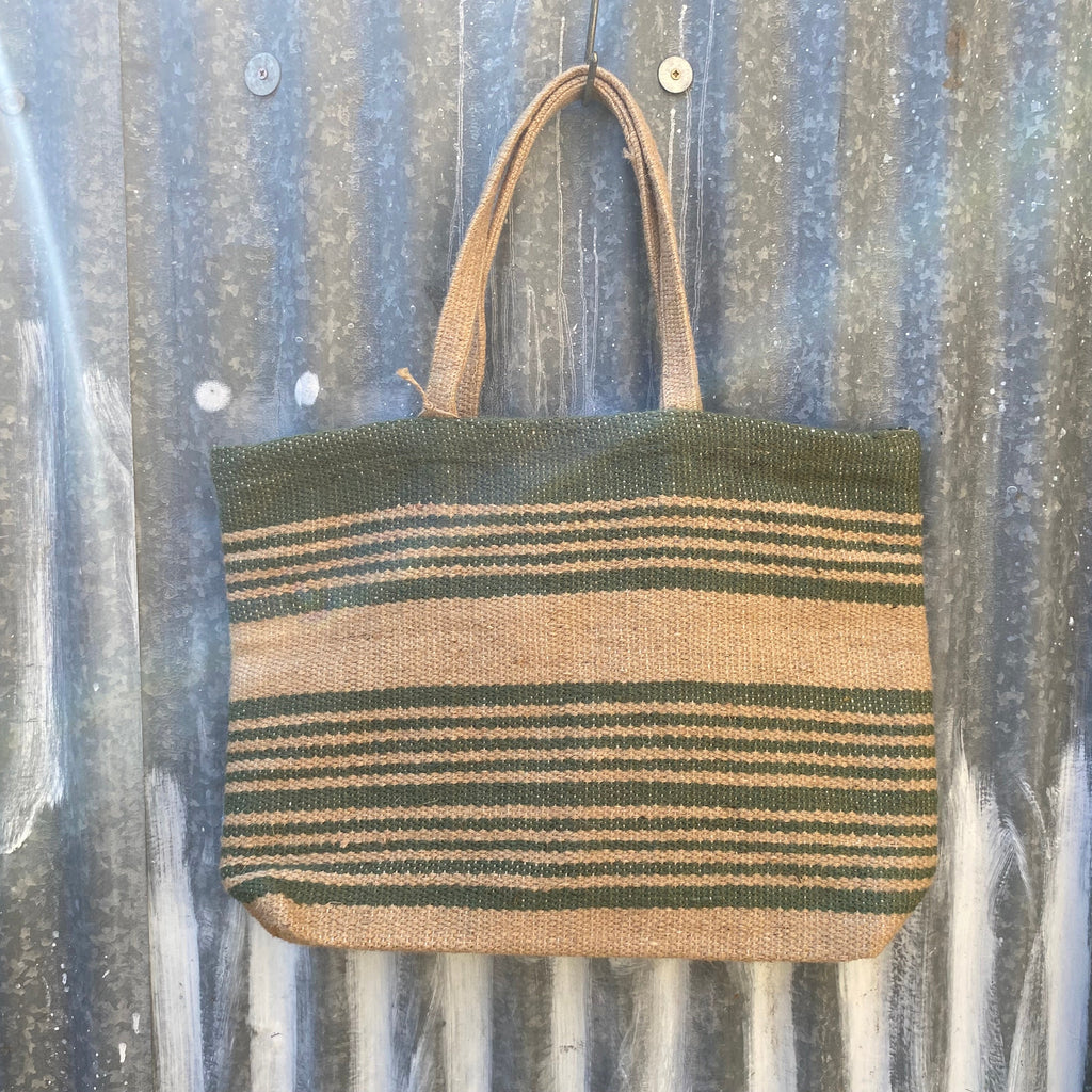 A woven jute 
tote bag with horizontal stripes in olive green and tan, hanging on a rusty corrugated metal wall.