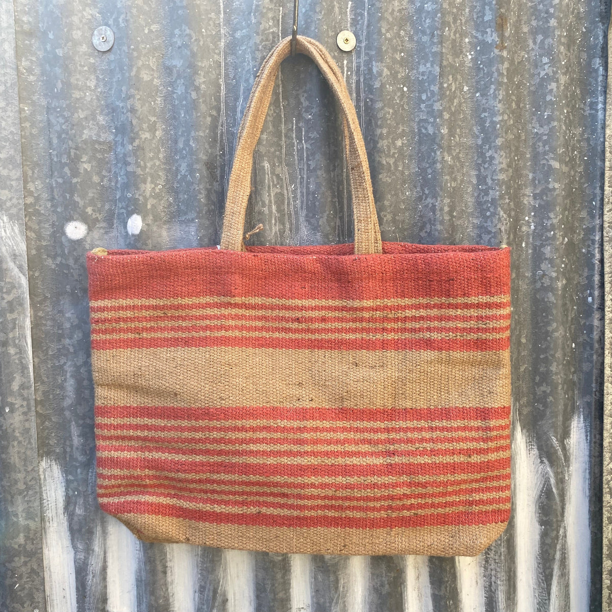 A woven tote bag made from natural jute with horizontal pink stripes, hanging against a corrugated metal wall.