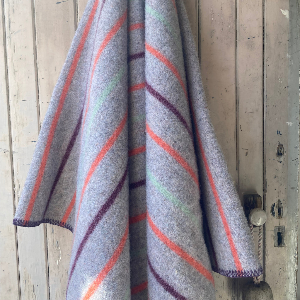 A gray wool blanket with diagonal stripes in orange, purple, and mint green, hanging on a rustic wooden door.