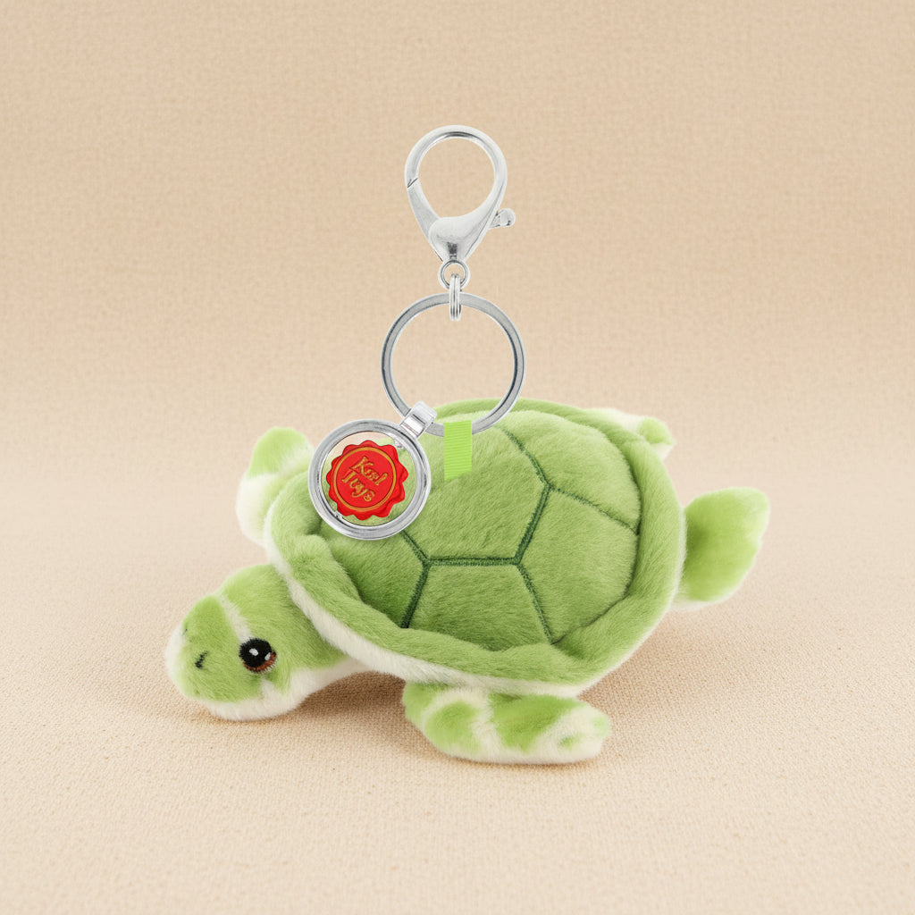 A plush green turtle keychain with a silver metal clasp and a round silver tag featuring a red seal, shown on a plain white background.