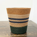 A woven basket with horizontal stripes in brown, tan, navy blue, and dark green, sitting on a rustic wooden table against a white wall.