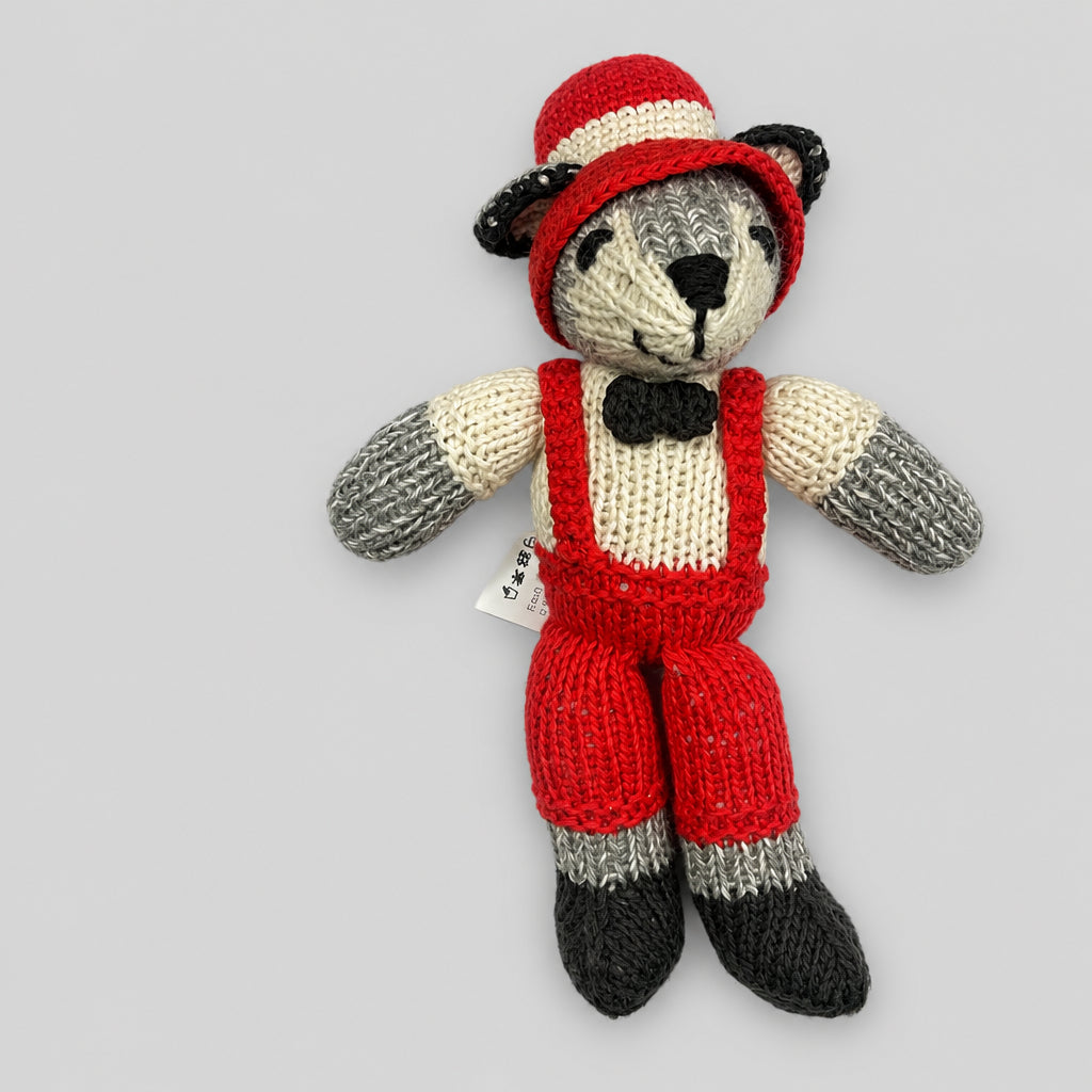 A hand-knitted wolf doll wearing red overalls, a red and cream striped hat, and a black bow tie, photographed against a neutral light gray background.