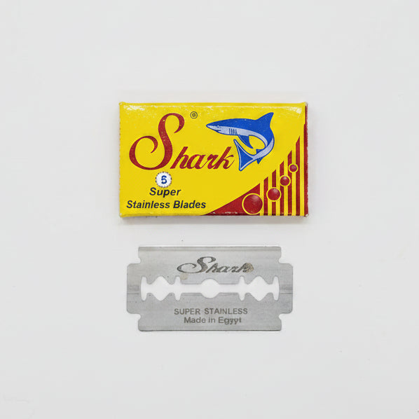 A pack of razor blades with a yellow and red label, indicating the brand 'Shark', with a single blade displayed in front of the packaging.