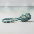 A handwoven straw rattle with a bell, featuring a green and natural-colored pattern.