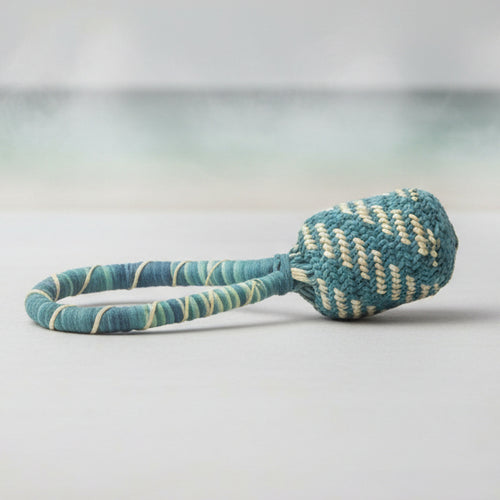 A handwoven straw rattle with a bell, featuring a green and natural-colored pattern.
