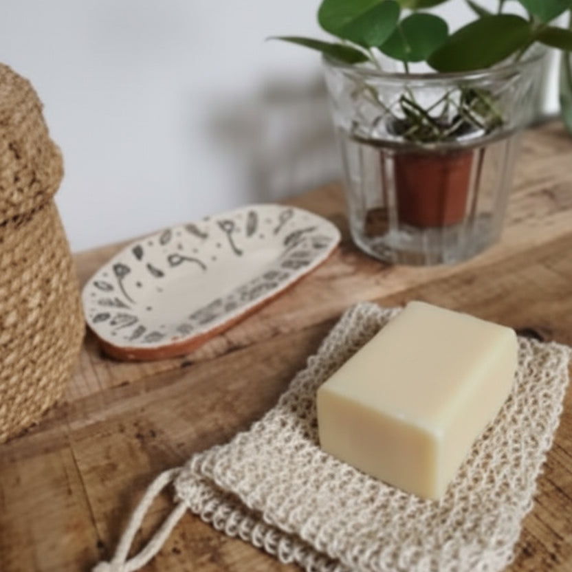 A bar of soap resting on a beige sisal mesh sack with a decorative vase and potted plant in the background, placed on a wooden surface.